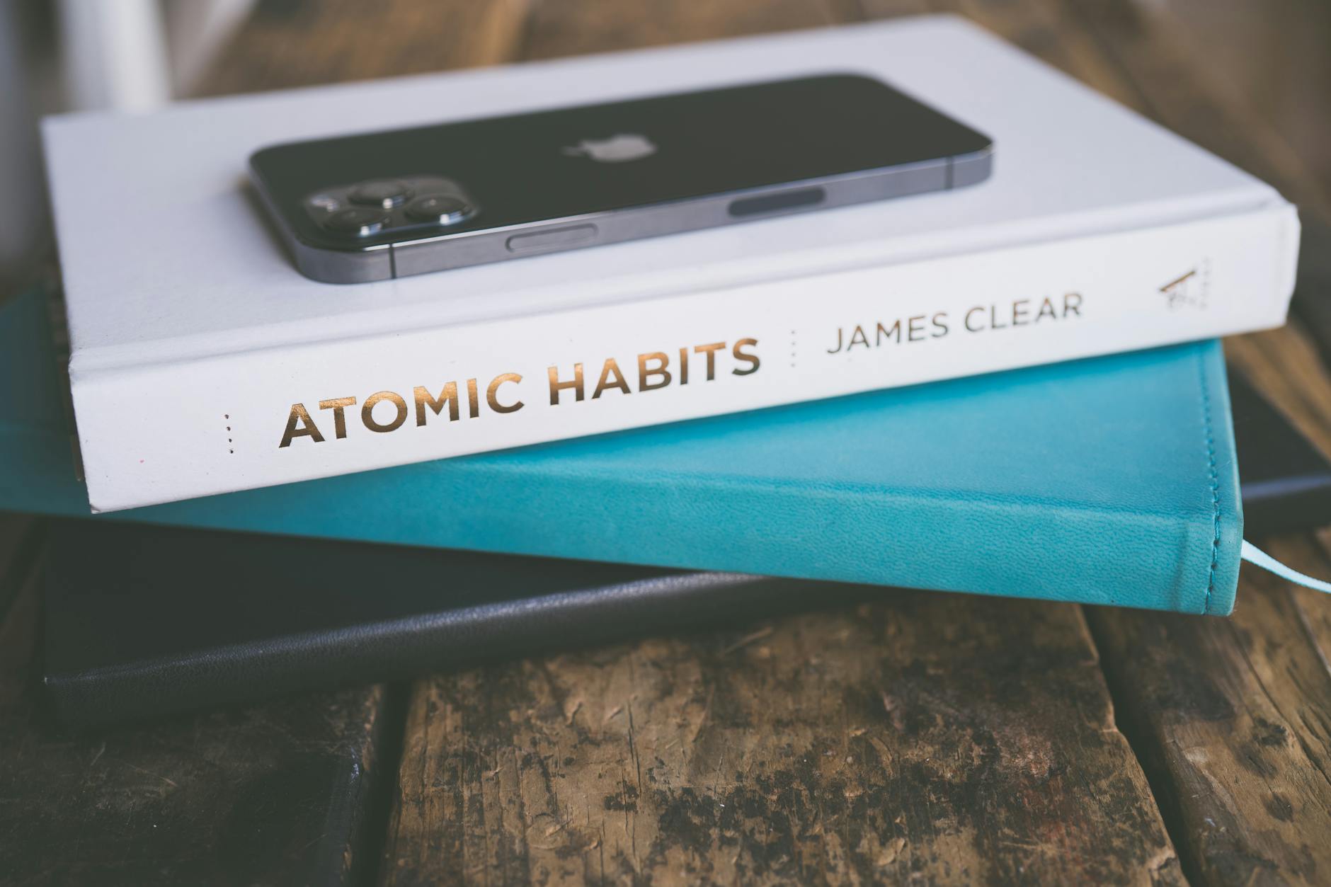 Close-up of a smartphone on a book stack, featuring 'Atomic Habits' by James Clear on a wooden su... (Photo by Jessica Lewis 🦋 thepaintedsquare on Pexels)
