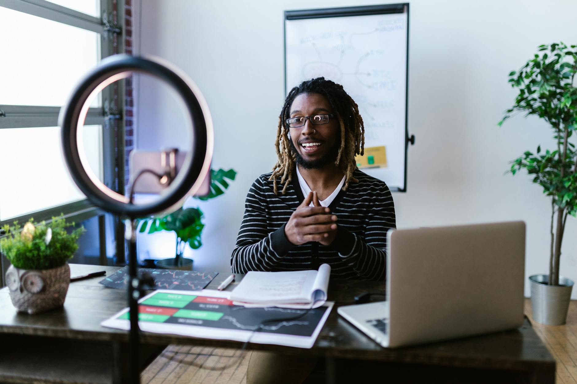 Young man with dreadlocks creating a video blog using smartphone and ring light in home office. (Photo by RDNE Stock project on Pexels)